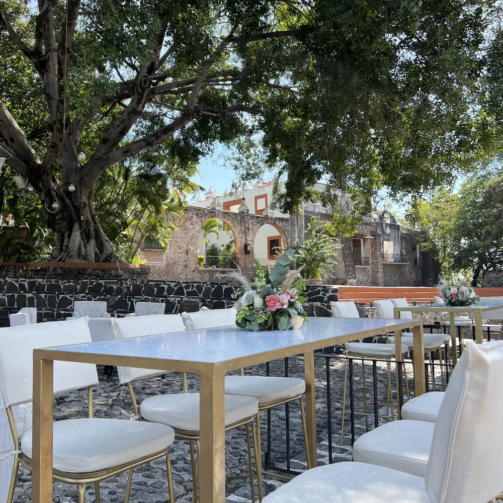 Mesas altas con base dorada y sillas blancas de AP Eventos bajo un árbol, con las ruinas de la hacienda al fondo