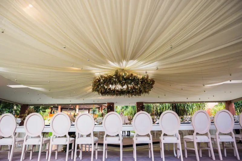 Banquete de boda bajo carpa drapeada con sillas blancas de respaldo ovalado