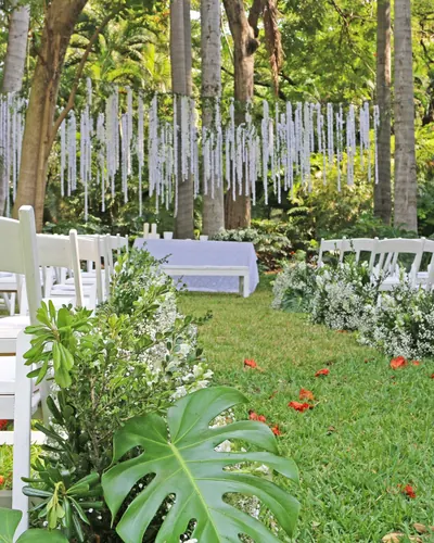 Pasillo de ceremonia en un jardín tropical, con sillas plegables blancas y arreglos de flores blancas en el suelo