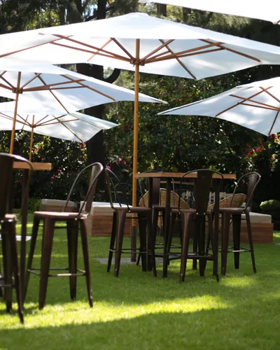 Periqueras de madera con bancos altos metálicos bajo sombrillas blancas en un jardín, montadas para un coctel