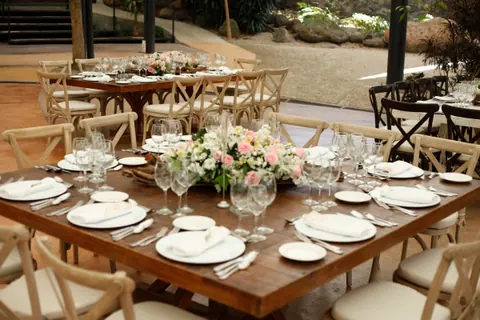 Mesa cuadrada de madera puesta con copas, platos y un centro de rosas, rodeada de sillas Crossback claras, con más mesas al fondo
