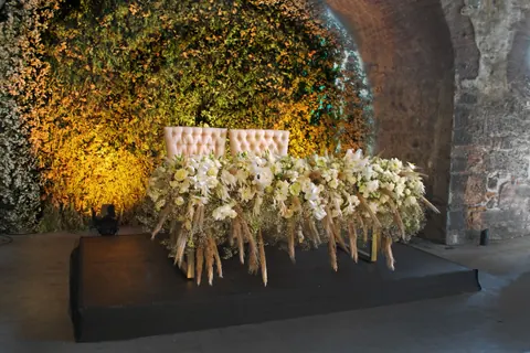 Mesa de novios con flores blancas y sillones rosados en una bóveda de piedra con muro verde iluminado
