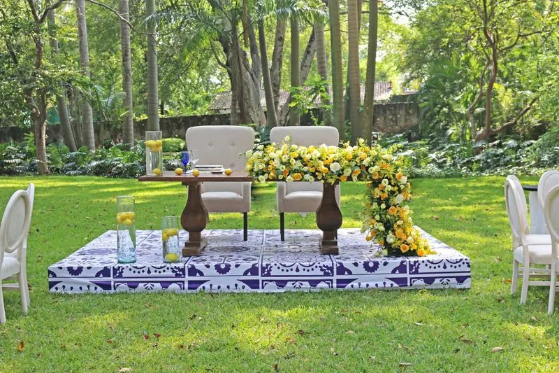 Mesa de novios con sillones capitonados y flores amarillas, sobre tarima con estampado azul y blanco en un jardín