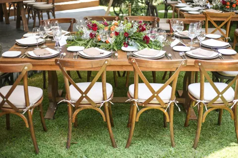 Mesa de madera puesta con un centro largo de flores rojas y naranjas, rodeada de sillas Crossback de madera con cojín claro, sobre pasto