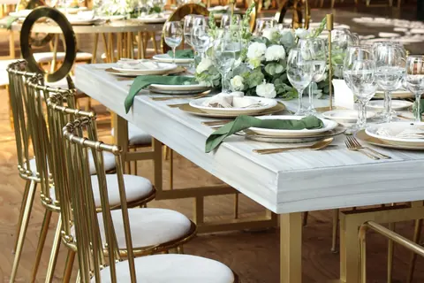 Mesa de cubierta blanca con sillas doradas de respaldo de barrotes bajo una carpa, con más mesas al fondo