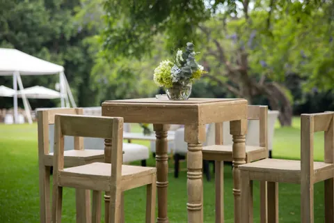 Periquera de madera rústica con bancos altos en un jardín, con una carpa blanca al fondo