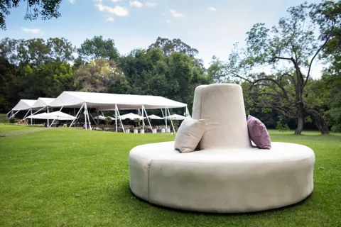 Puff redondo blanco con cojines en un jardín amplio, con carpas blancas al fondo