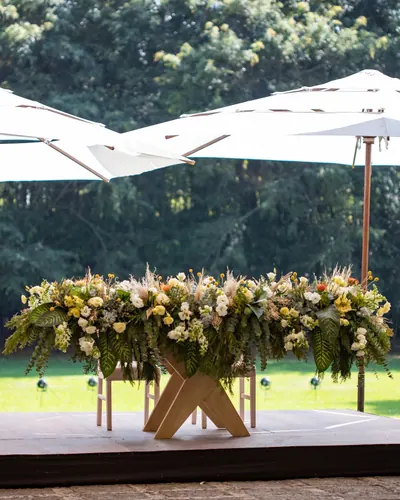 Mesa de novios de madera con base en cruz cubierta de flores, sobre tarima y bajo dos sombrillas blancas