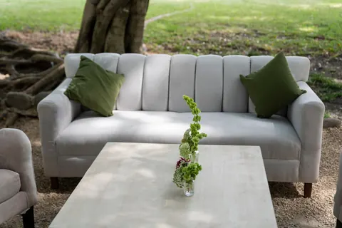 Sala lounge con sofá blanco de respaldo acanalado, cojines verdes y mesa de centro clara, sobre grava bajo un árbol