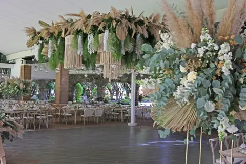 Salón abierto con una gran instalación colgante de pampas y follaje sobre la pista y mesas montadas alrededor