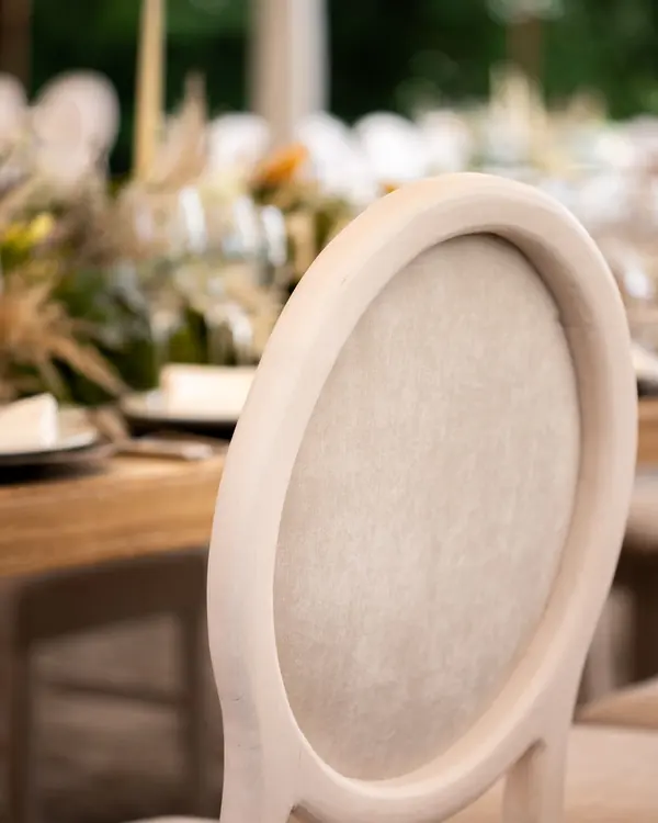 Detalle del respaldo ovalado tapizado de una silla de madera clara, con una mesa puesta desenfocada detrás