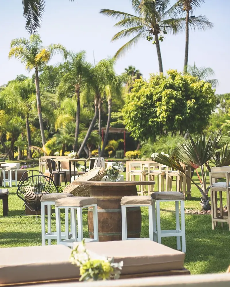 Zona de coctel en un jardín con palmeras: periquera de barril con bancos altos blancos, mesas altas de madera, sillas tejidas y bancas bajas con cojín